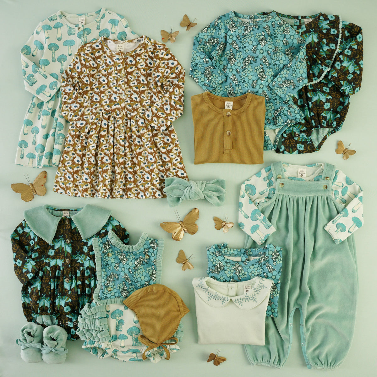Flat lay of a kid clothing collection on a mint green background. Items include long-sleeve tops and dresses in fungi and daisy prints, floral bubbles and bodysuits in teal and dark butterfly print, a mustard henley top, a sage velour jumpsuit, flight hat, and booties, a velour knot headband, and embroidered collar tops — accented with decorative gold butterfly ornaments.