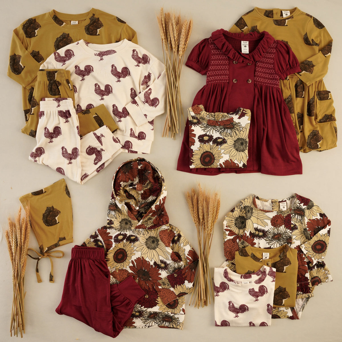 Flat lay of baby and kids clothing in autumn colors on a beige background with bundles of dried wheat. Includes a mustard squirrel-print sweatshirt, pants, dress, and elf hat; a cream rooster-print sweatshirt, joggers, and tee; a burgundy short-sleeve dress with a ruffled collar, smocking, and double-breasted buttons; and a cream sunflower-print hoodie and bubble next to with burgundy pants.