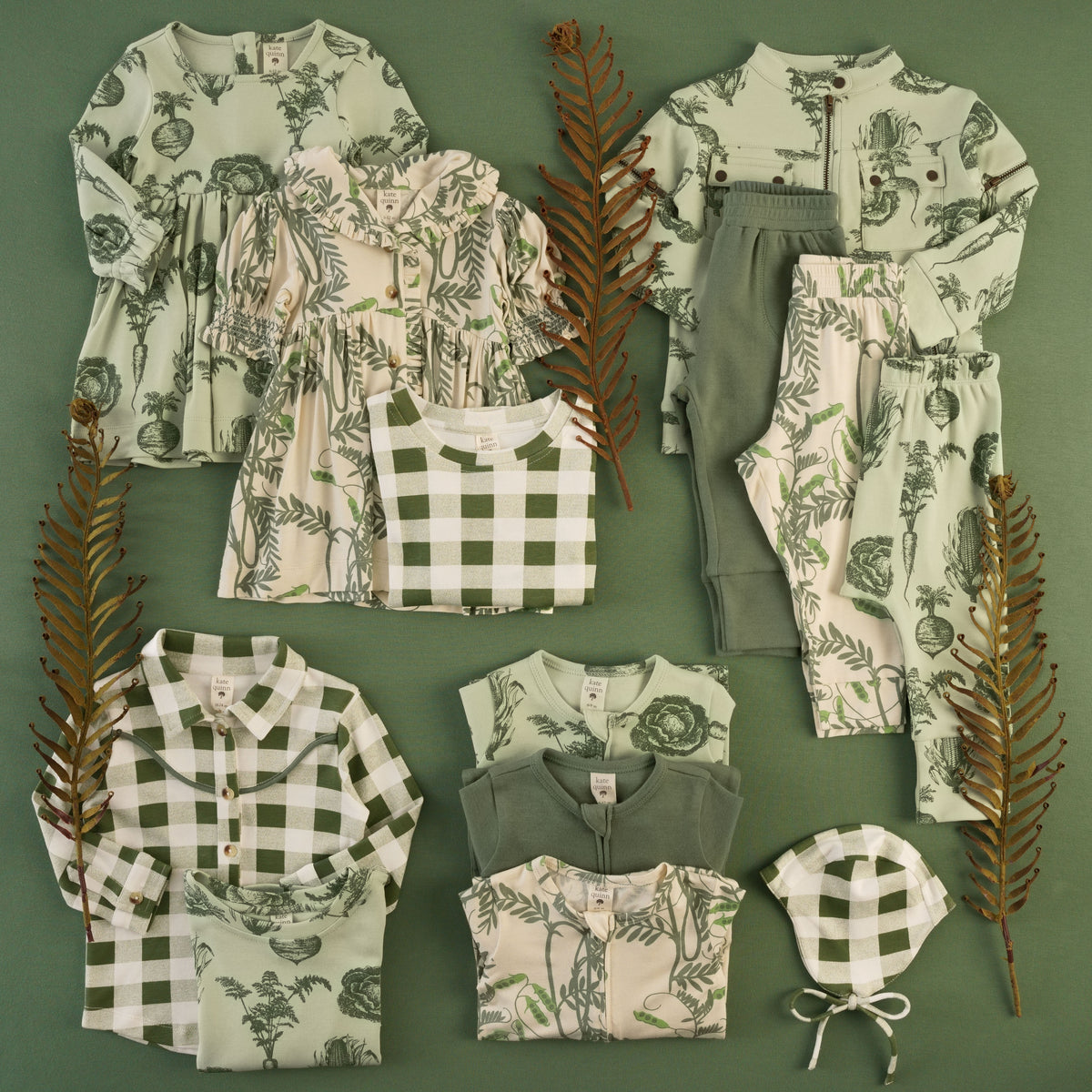 Flat lay of baby and kids clothing on a sage green background with dried fern fronds. Includes pieces in a sage vegetable-garden print with beets, carrots, cabbage, and corn, such as a long-sleeve dress, zip jacket with snap pockets, and bottoms; a cream pea-vine print dress with a ruffled collar and smocked sleeves; green and white gingham tops, a collared button-up shirt, and a flight hat; and solid green joggers and zip footies.