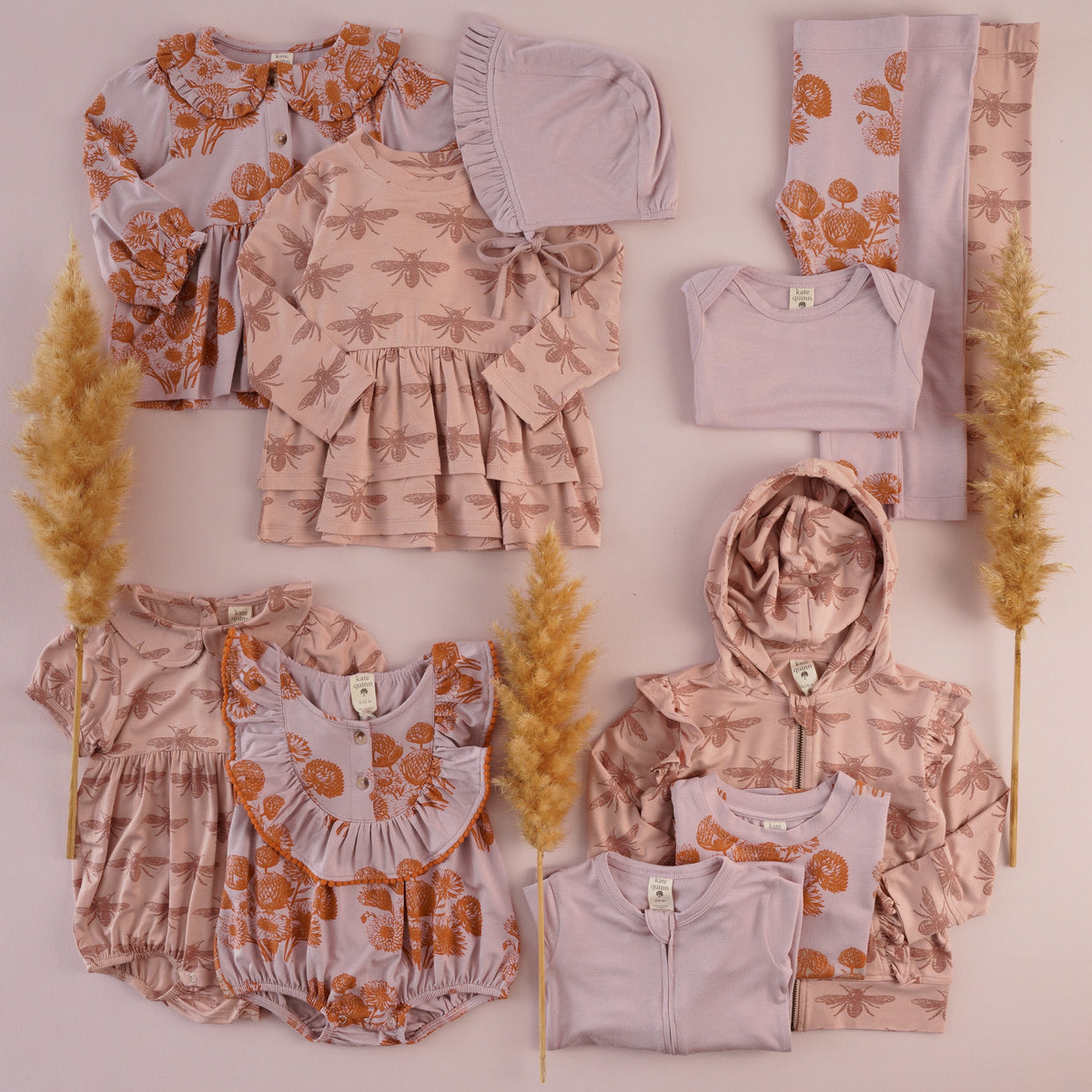 Flat lay of baby and kids clothing in soft pink and lavender on a blush background with pampas grass. Includes a dusty pink bee-print tiered dress, hooded zip jacket with ruffled shoulders, and Peter Pan collar bubble; lavender pieces with a rust-orange dahlia print, such as a ruffle-collar cardigan, a pom-pom trimmed ruffle-bib bubble and leggings; and a solid lavender ruffled bonnet, bodysuit, and zip footie.