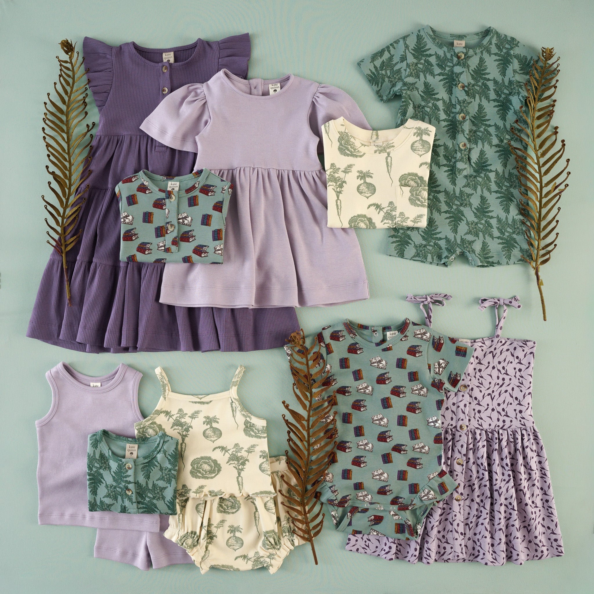 Flat lay of children's clothing in soft lavender, sage green, and cream tones, featuring dresses, rompers, bodysuits, and matching separates with botanical and storybook-inspired prints.