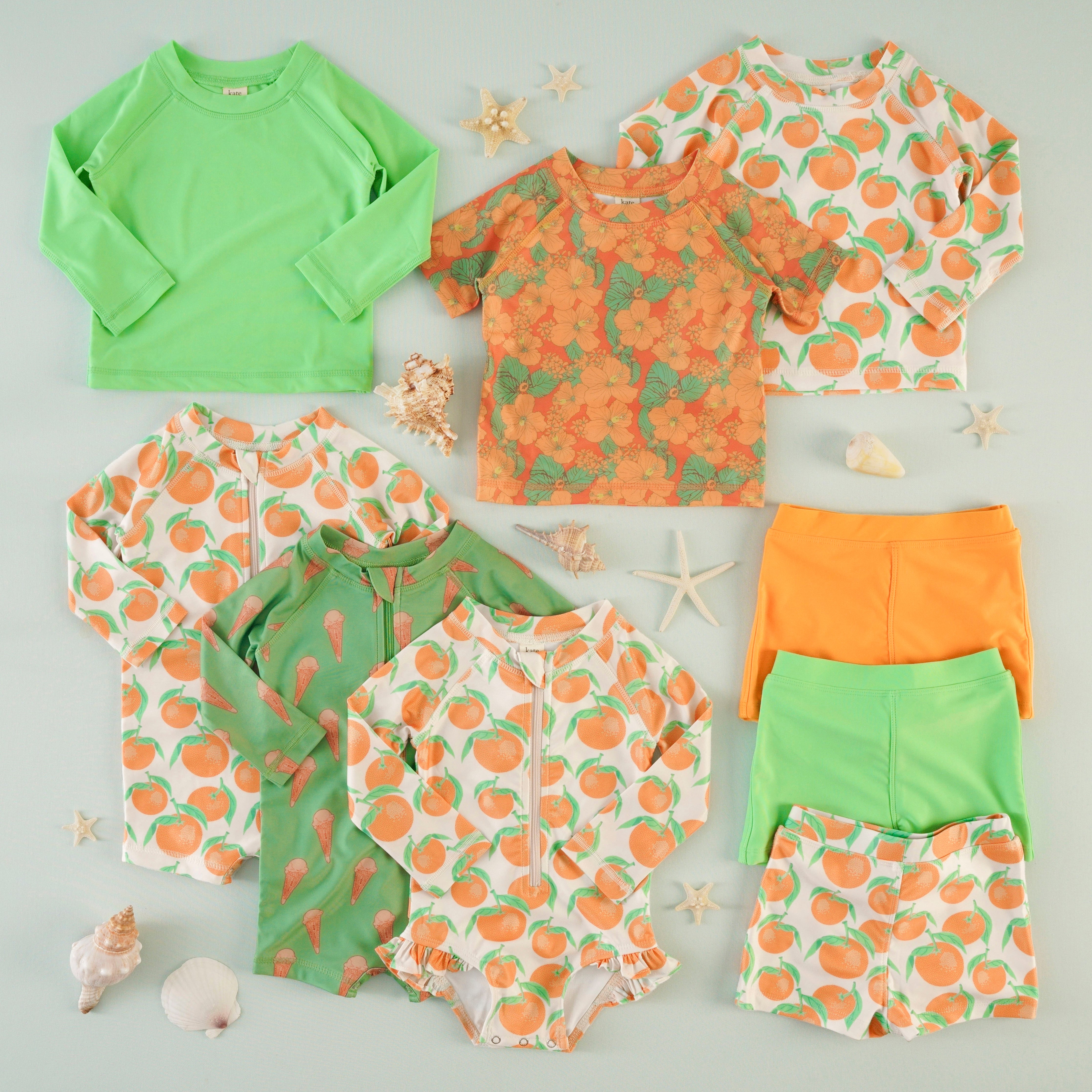 A styled flatlay of a coordinating children's swimwear collection on a light blue background. Pieces include long-sleeve rash guards, zip-front one-piece swimsuits, and swim shorts. The color palette is bright orange and lime green, with three prints: an all-over orange clementine print, an ice cream cone print, an orange hibiscus print, and a solid lime green. Seashells and starfish are scattered throughout as props.