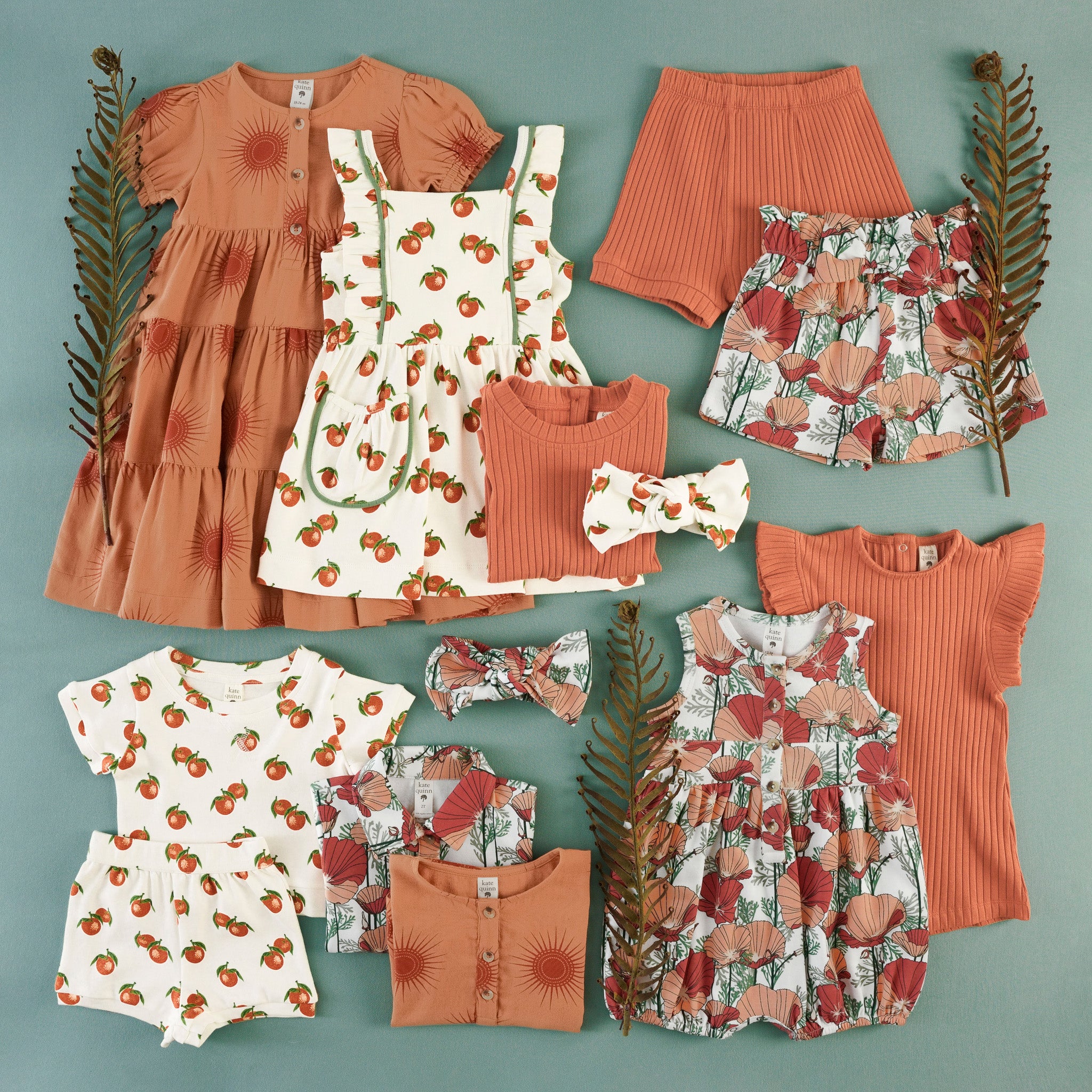 Flat lay of children's clothing in orange and rust tones: a sunburst-print tiered dress, a clementine-print ruffle-strap pinafore dress with pockets, a rust ribbed top, ribbed shorts, a poppy floral print collar top, clementine-print shorts and top set, poppy floral shortie, a ribbed ruffle-sleeve shortie, poppy floral front bow shorts, and knotted bow headbands in clementine and poppy prints.