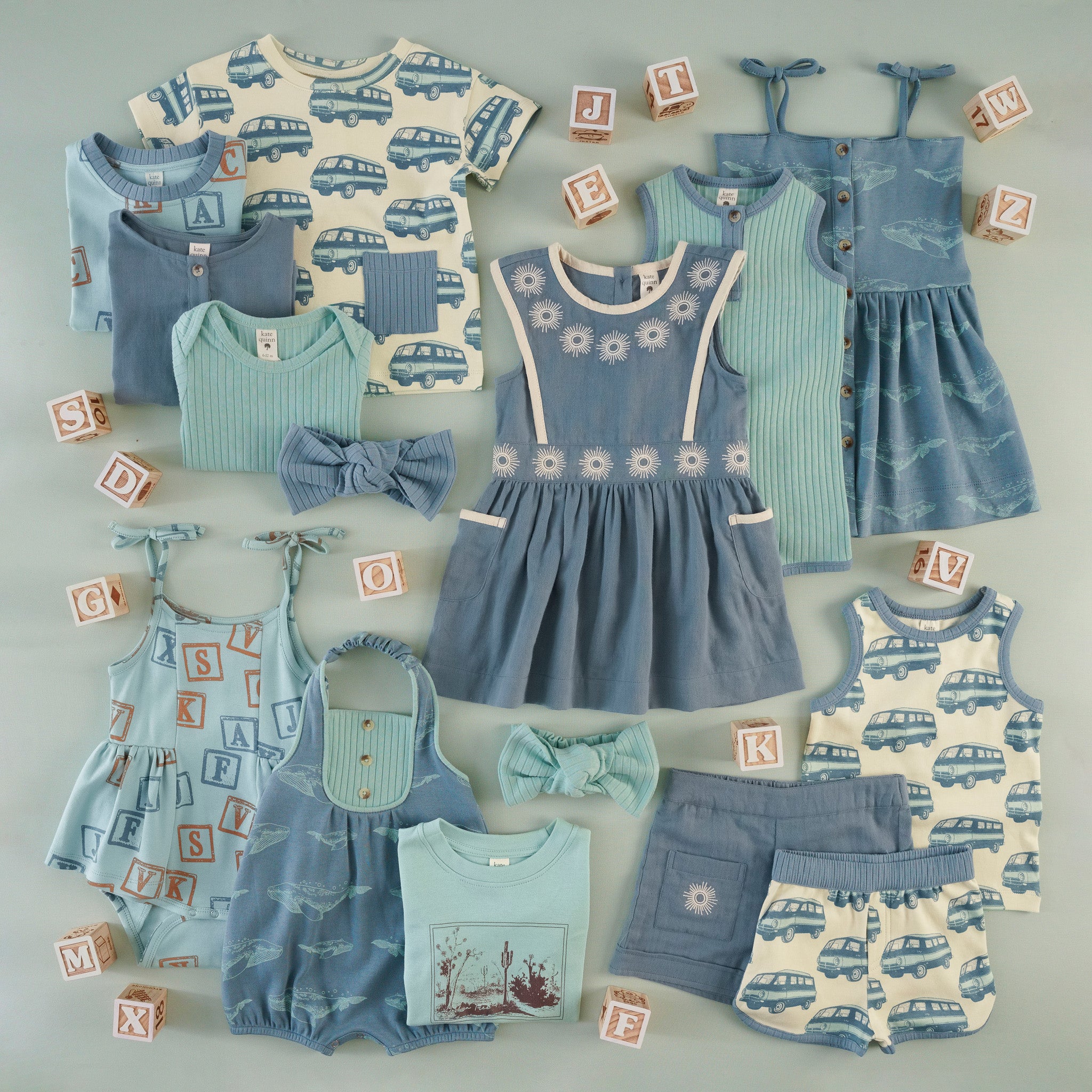 Flat lay of children's clothing in blue and aqua tones: a beach van print top, ribbed ringer top, aqua bodysuit, sunburst-embroidered pinafore dress, aqua tank shortie, whale-print dress, alphabet block-print bubble, whale print shortie, a desert landscape screen print tee, sunburst-pocket shorts, beach van print shorts and tank set, and bow headbands, with wooden alphabet blocks as prop.