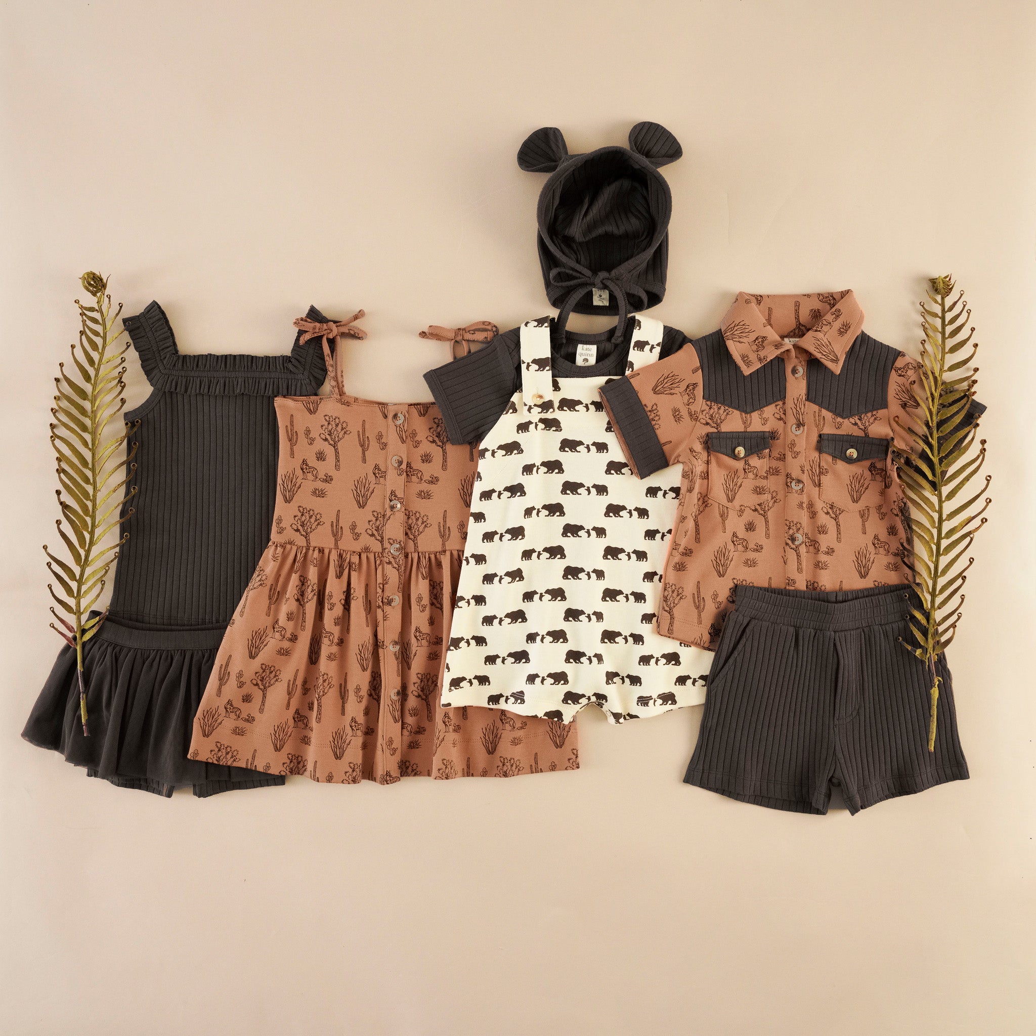 Flat lay of children's clothing, a chocolate ribbed ruffle tank and tulle shorts, a rust button-front dress with a desert landscape print, a cream and brown shortie with bear print, a chocolate rib bodysuit with matching bear hat, a rust western-style button-up shirt with a desert landscape print and chocolate contrast trim, and chocolate ribbed shorts.