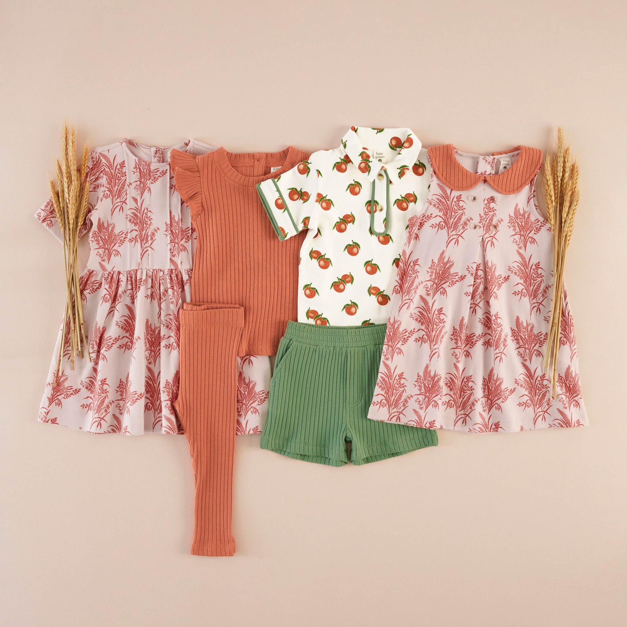 Flat lay of children's clothing in mauve and rust tones: two wheat/grass print dresses (one button-back, one with a Peter Pan collar), a rust ruffle-sleeve top, a citrus-print collared polo shirt, ribbed rust leggings, and green ribbed shorts, styled with dried wheat stalks.