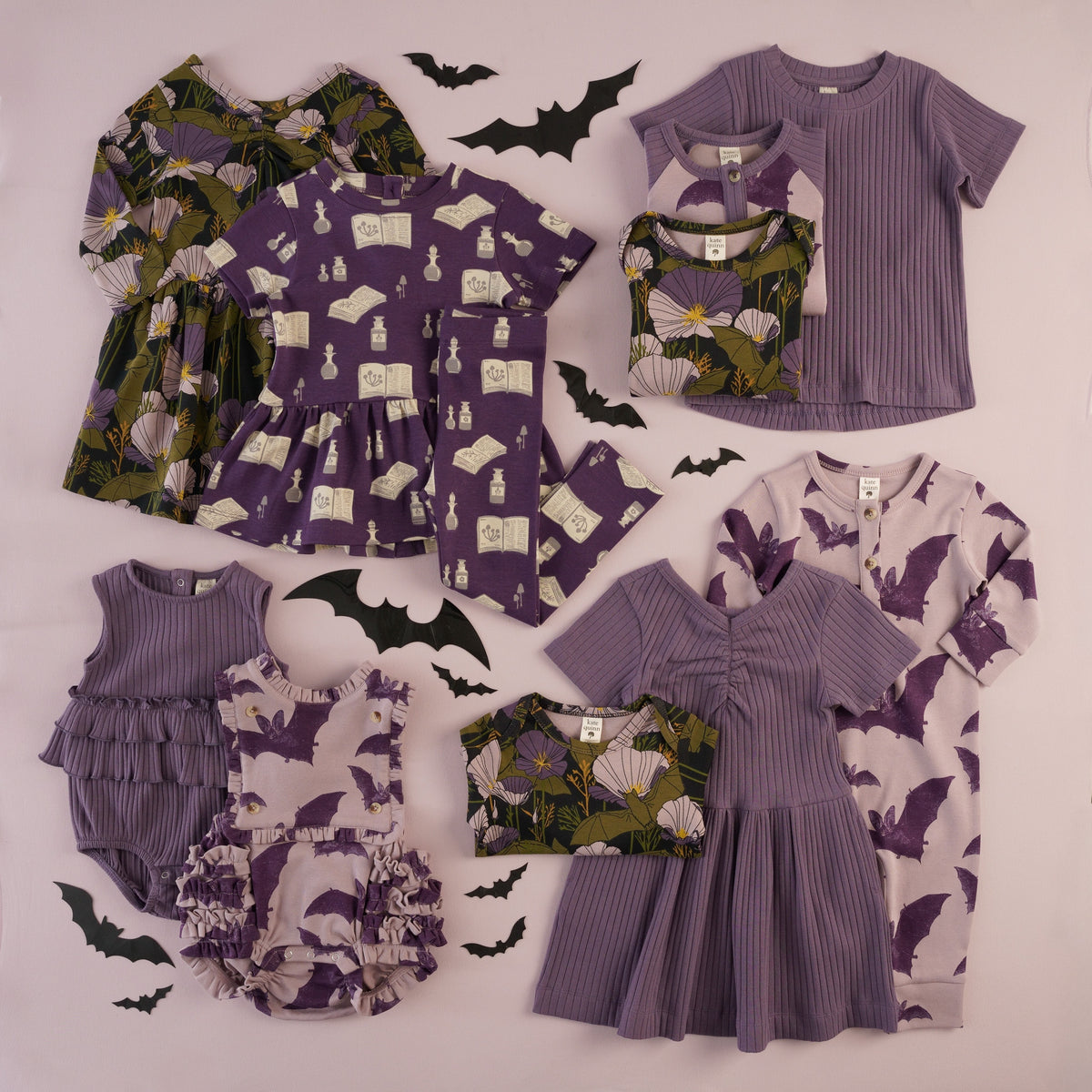 Flat lay of purple Halloween-themed kids' clothing on a lavender background, including a poppy + bat dress, a witchy potion-and-spellbook print top with matching leggings, a bat-print jumpsuit, ribbed purple tops, ruffled bubbles, and dresses, styled with black paper bat cutouts.