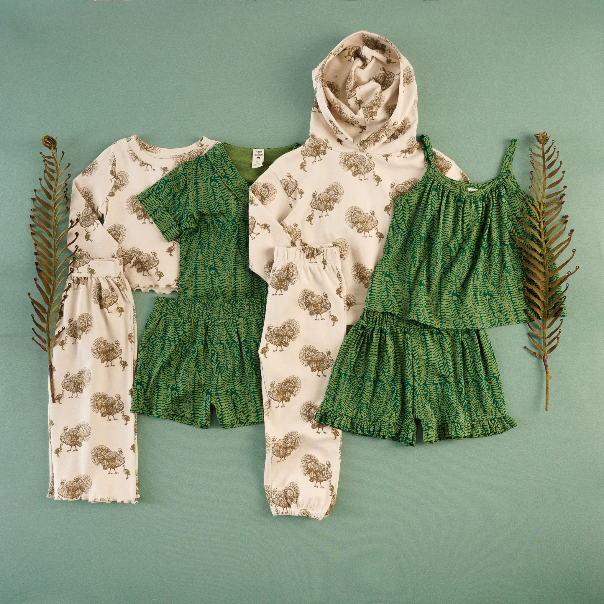 Flat lay of children's clothing in a turkey print and green fern print collection on a seafoam green background, styled with dried fern fronds. Includes a cream turkey-print long-sleeve top and matching pants, a cream turkey-print hoodie, green fern-print short-sleeve top and shorts set, and a green fern-print camisole top with ruffled shorts.