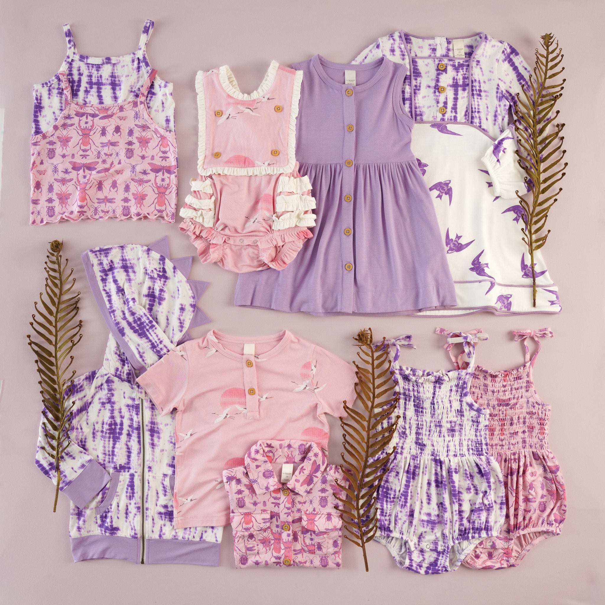 Flat lay of children's clothing in shibori, pink and purple insect print, cranes, and sparrow prints on a pale pink background, styled with dried fern fronds. Includes a pink insect-print tank top, pink crane-print ruffled bubble romper, solid lavender button-front dress, purple sparrow-print dress, shibori-dye dinosaur-spike hoodie and bonnet, pink crane-print tee, pink insect-print jacket, and shibori smocked rompers in purple and pink.