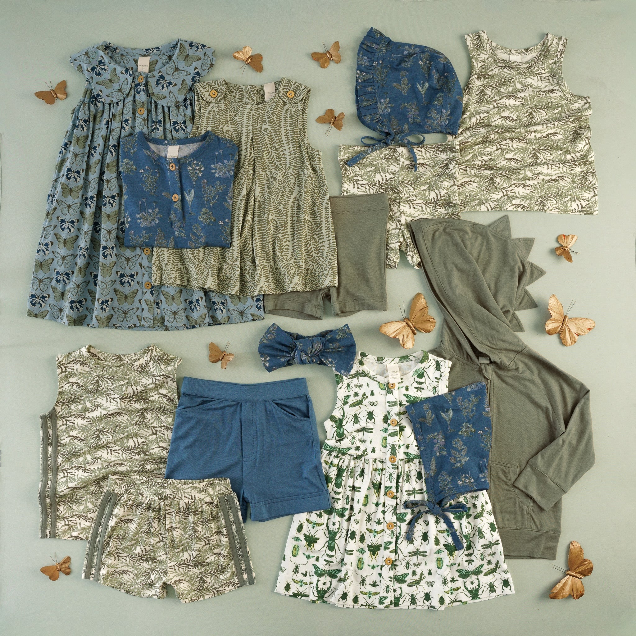 Flat lay of children's clothing in a butterfly, fern, and insect print collection on a sage green background, with butterfly decorations. Includes a blue butterfly-print dress, olive fern-print sets, tank top and bike set, blue floral henley shirt, blue bonnet with ruffle trim, blue shorts, olive shorts and dinosaur-spike hoodie, and a white and green insect-print dress.