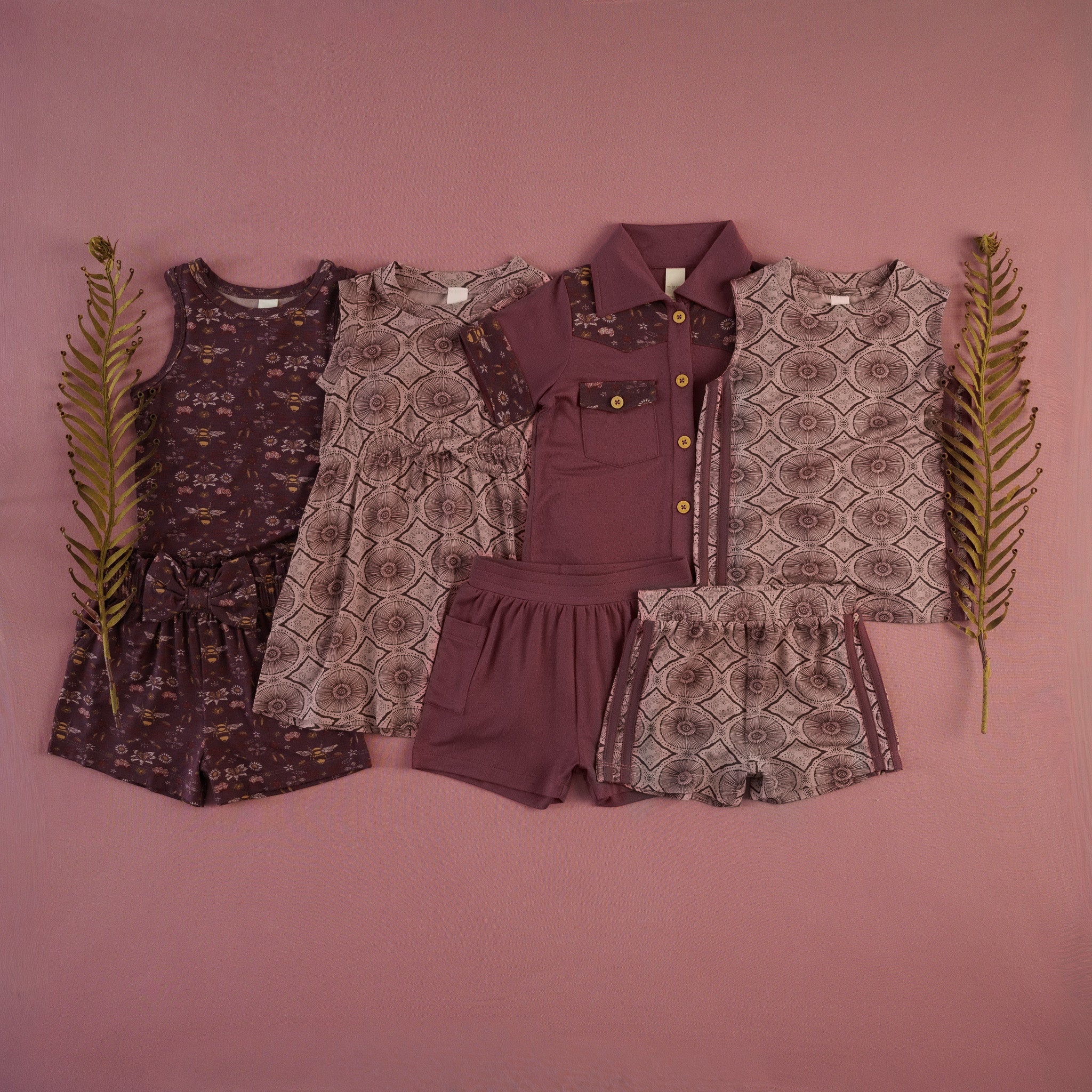 Flat lay of children's clothing in a mahogany bee floral and batik print collection on a dusty rose background, styled with dried fern fronds. Includes a mahogany bee-and-floral print short set with bow detail, a mauve batik-print dress, a solid mauve collared onesie with bee-floral trims, mahogany shorts, batik-print shorts with side stripes, and a batik-print sleeveless top.