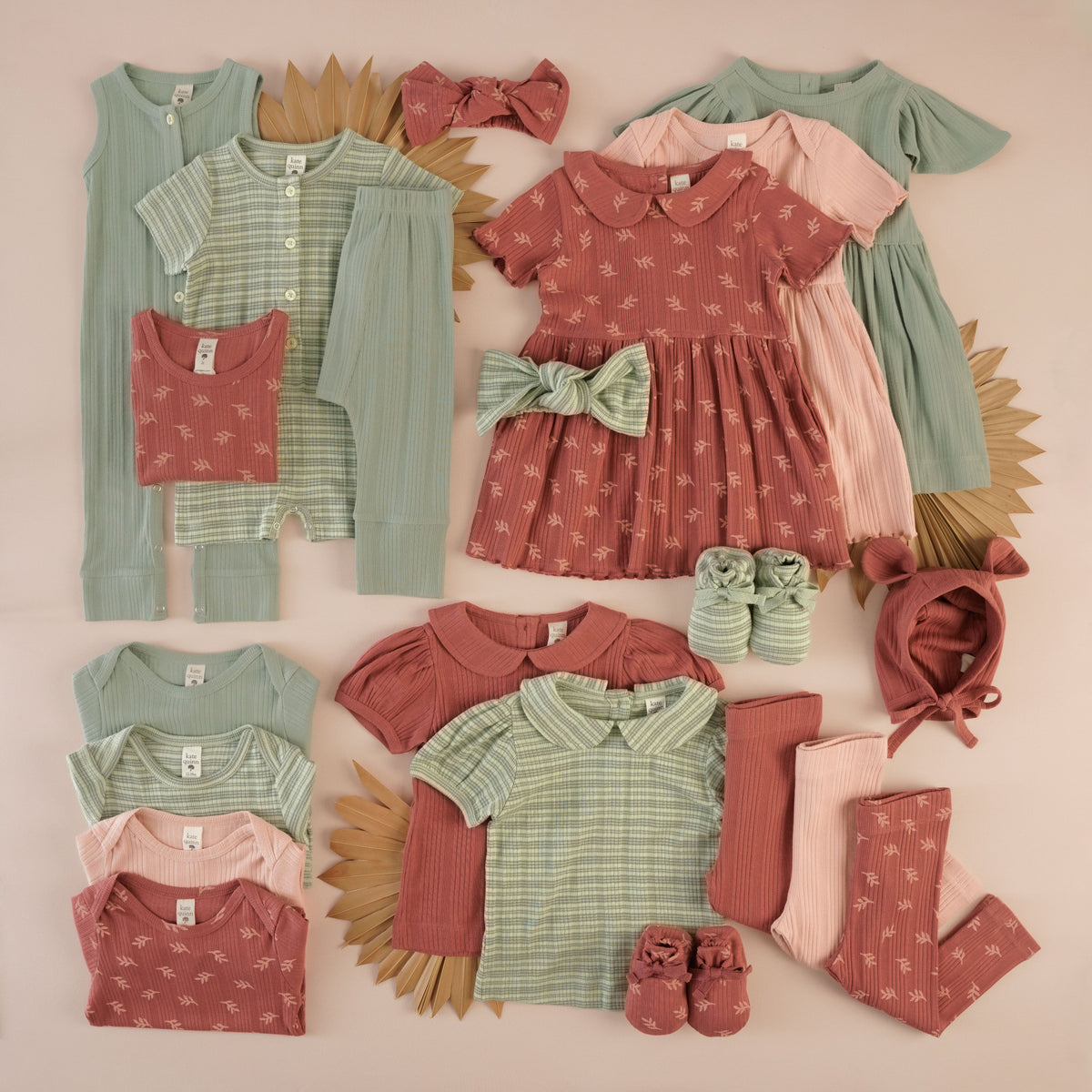 Flat lay of dusty rose leaf, sage green, green stripe print, and light pink kids' dresses, shortie, jumpsuit, tops, bodysuits and accessories with palm fan props.