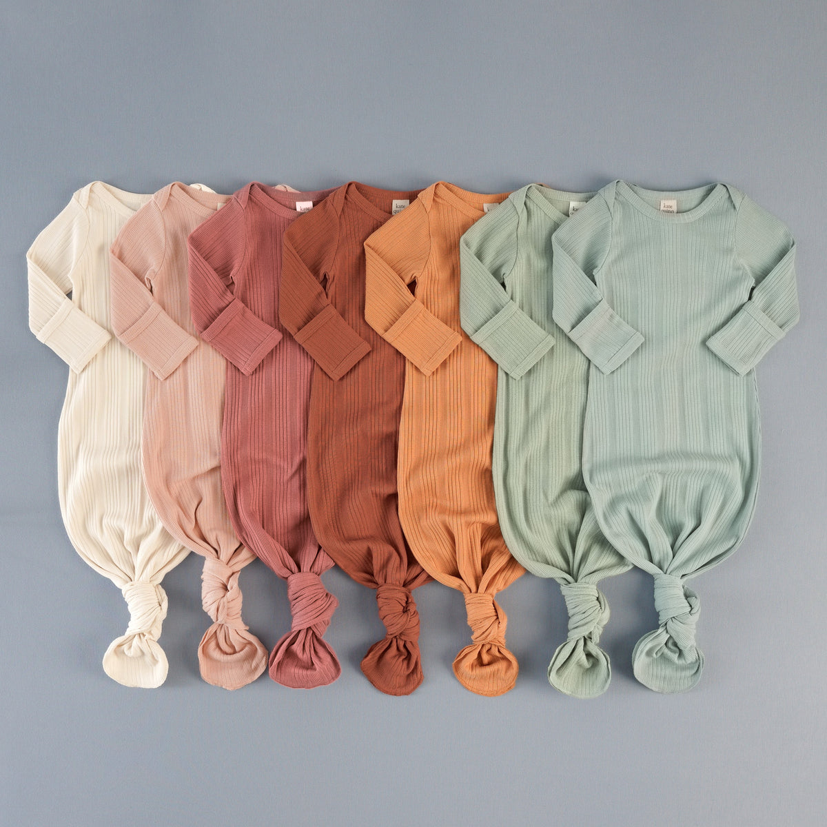 Flat lay of long-sleeve ribbed knotted gowns in a rainbow gradient of cream, pink, rust, orange, sage green, and blue shades.
