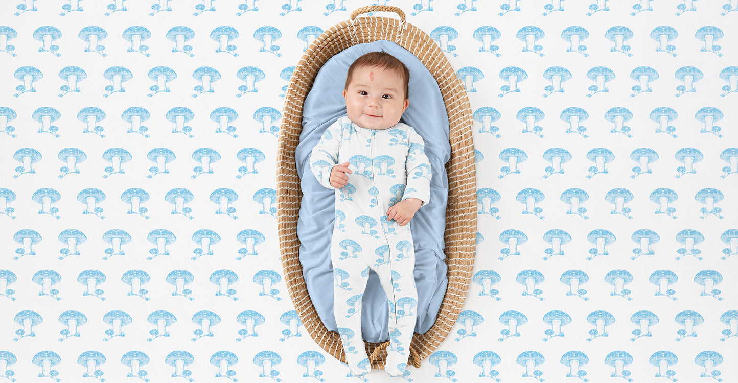 Baby smiling in a blue mushroom print zipper footie, lying in a woven bassinet with matching mushroom print background.