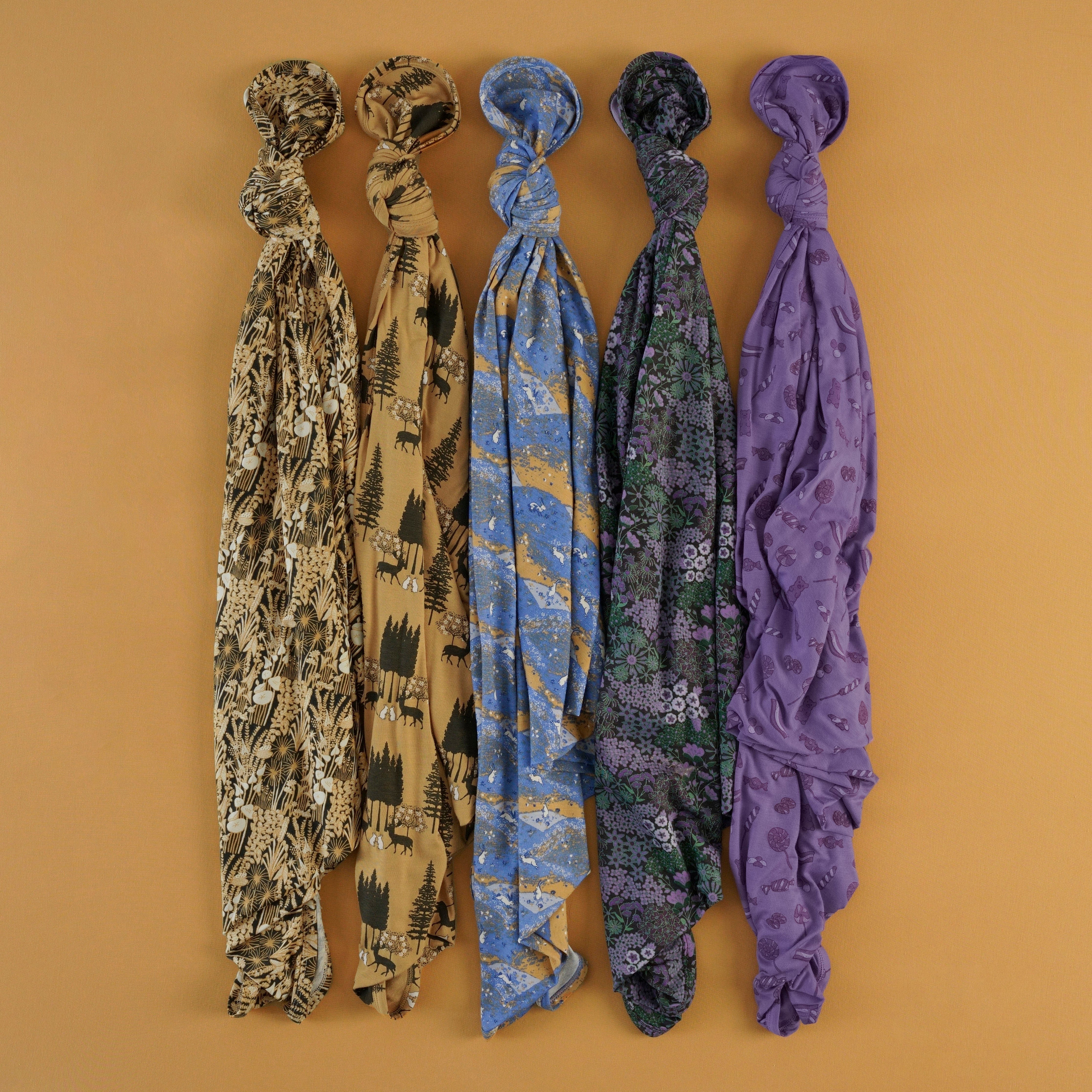 Five swaddle blankets twisted and hung vertically against a mustard orange background, showing five different prints from left to right: tan with a woodland foliage print, tan with a forest and forest animals print, blue with a mountain with a bunny print, navy with a purple and green floral print, and purple with a candies print.