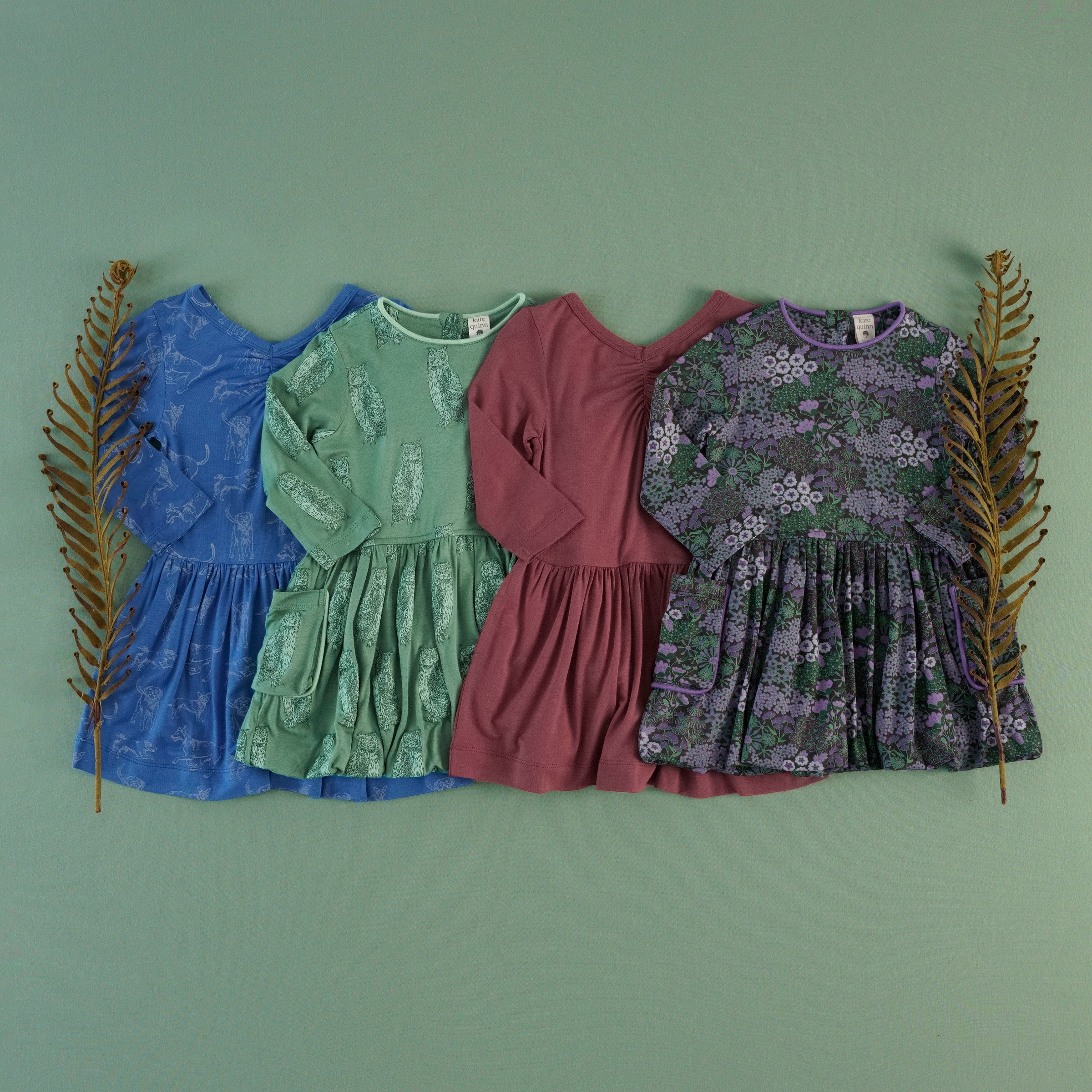 Four long-sleeve dresses lay flat and overlapping on a sage green background, flanked by two decorative dried fern branches, showing four different colors and prints from left to right: blue with a mutt print, sage green with an owl print, solid burgundy, and purple with a floral print.