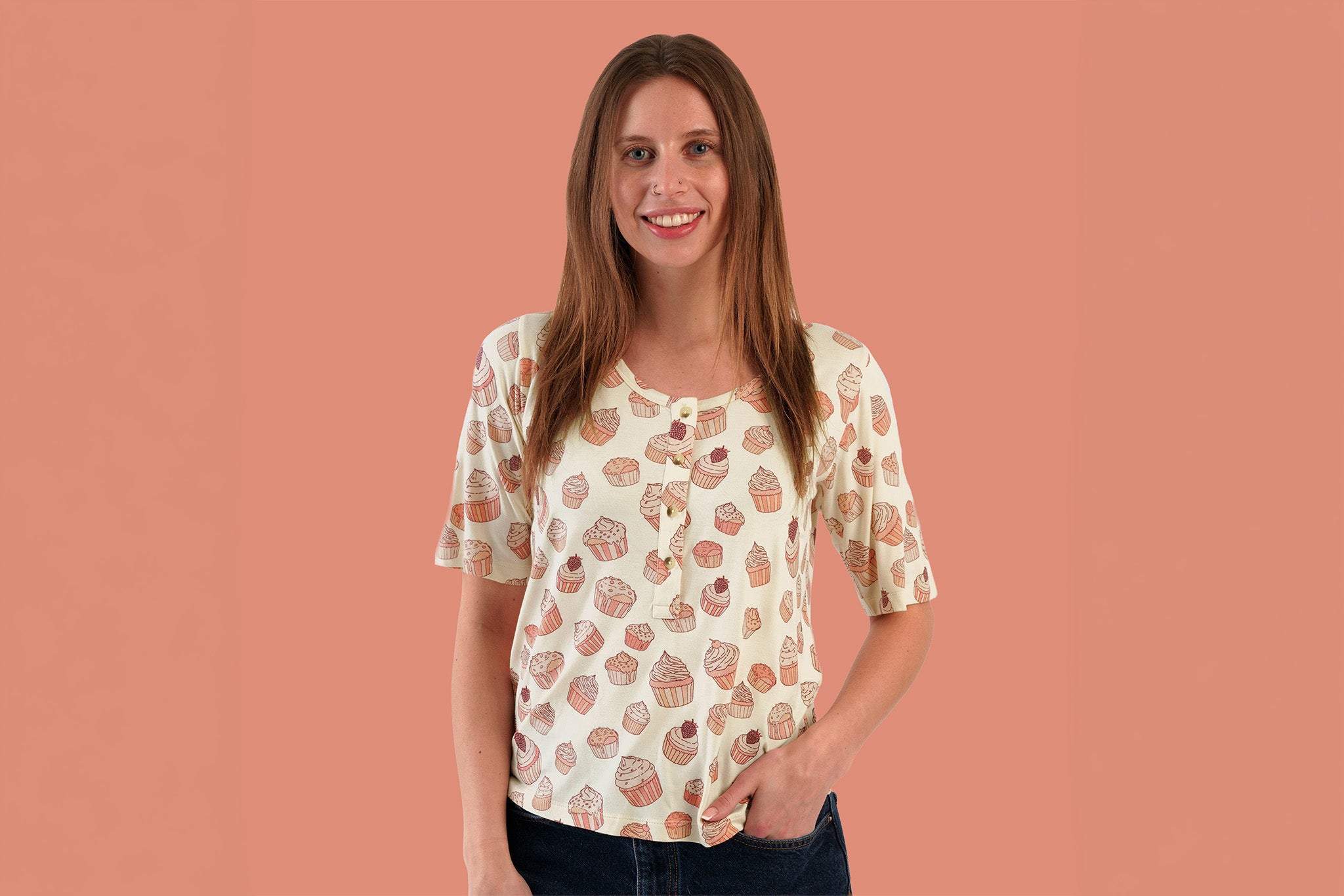 Woman modeling cream henley top with allover cupcake print in coral and pink tones, featuring short sleeves, scoop neckline, and button placket, worn with dark denim against a dusty coral background.