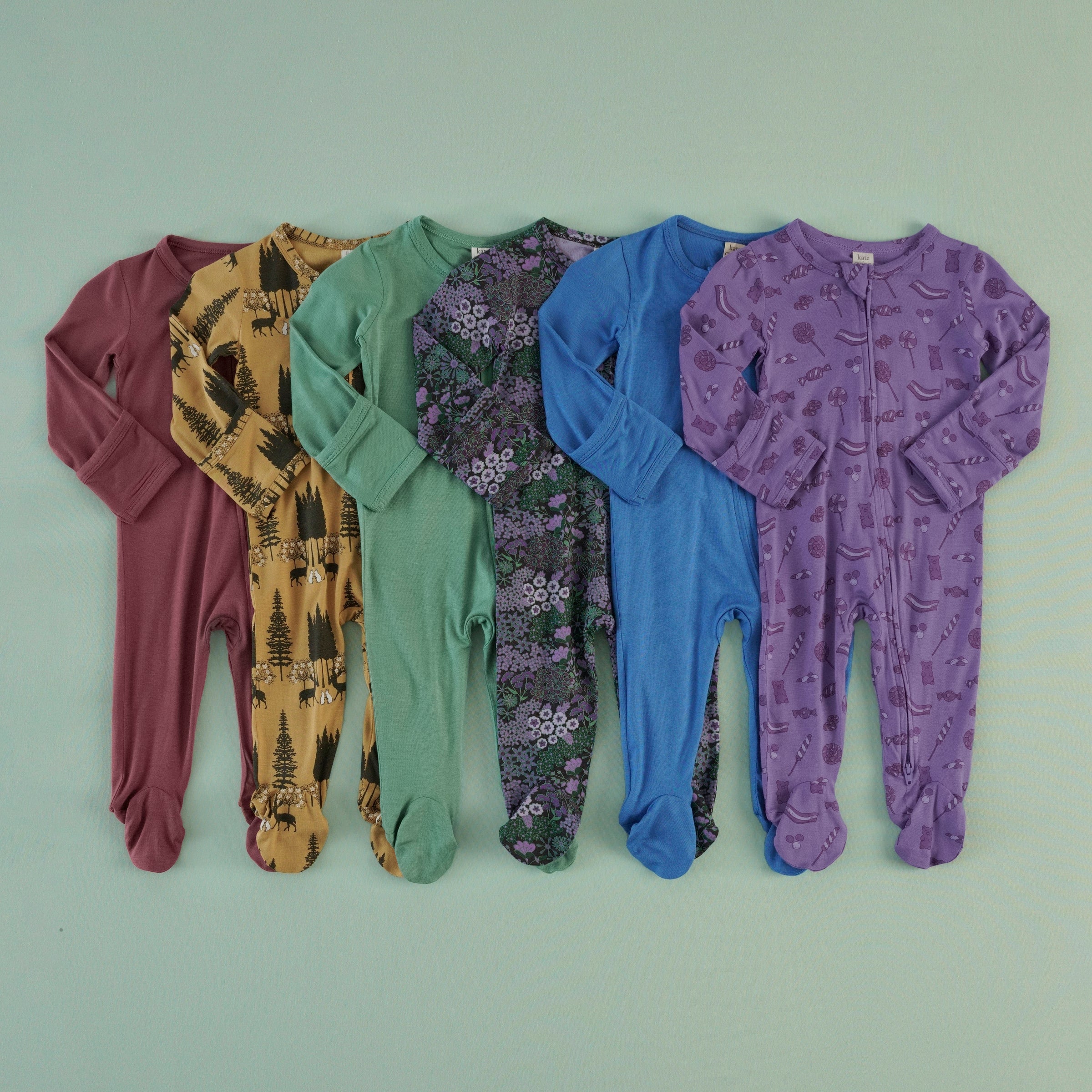 Six long-sleeve footies laid flat and overlapping on a sage green background, in the following colors and prints from left to right: solid burgundy, mustard yellow with a forest and forest animals print, solid sage green, purple with a dark floral print, solid blue, and purple with candies print.