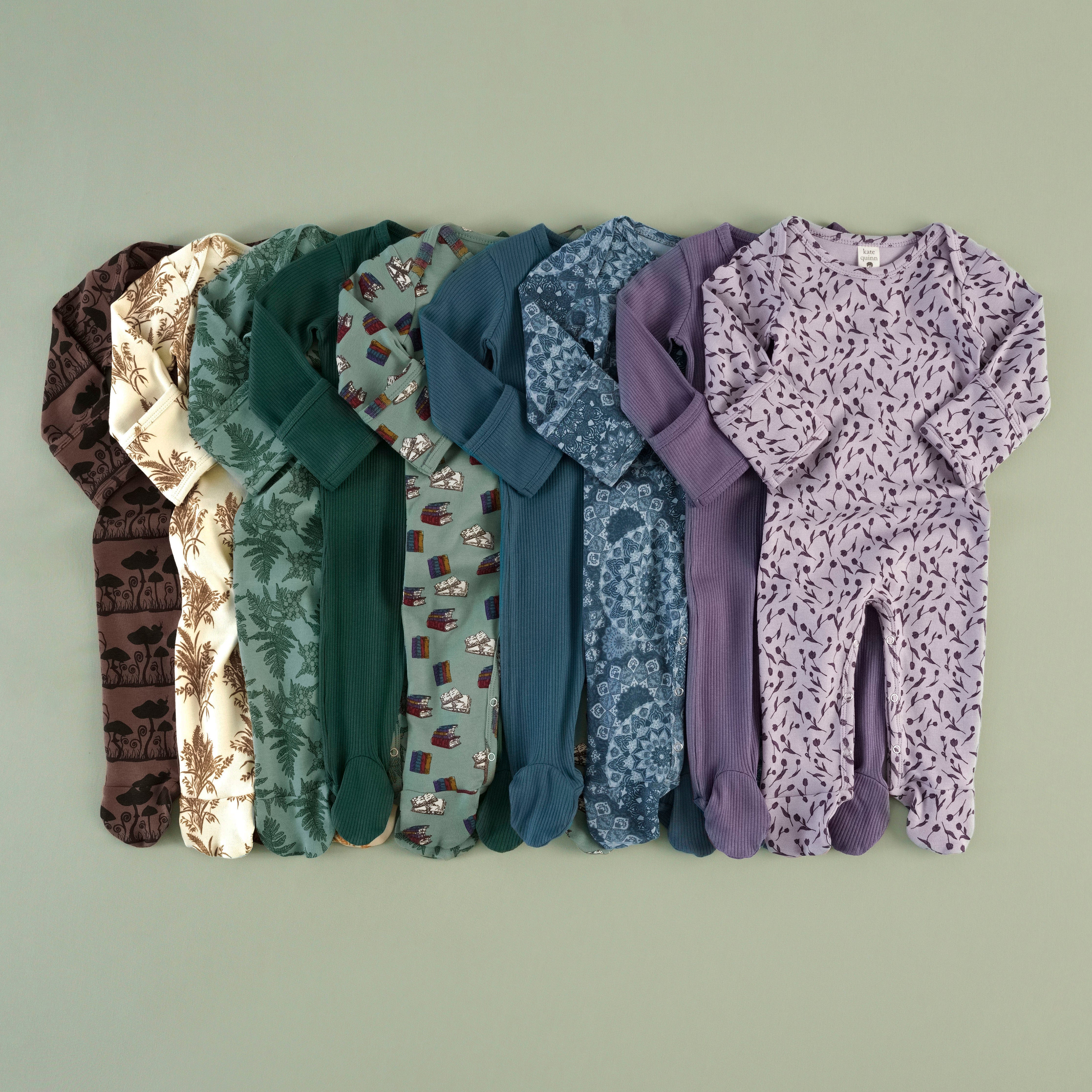 Row of 9 footies overlapping in a fan layout, in mushroom silhouette, wheat print, green fern, ribbed green, book print, ribbed blue, blue mandala print, ribbed purple, and purple-lavender tulip print.